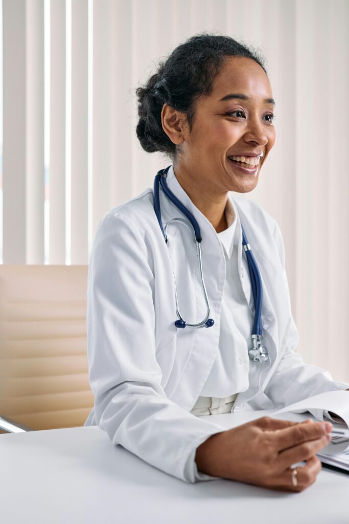Black female doctor smiling at desk with stethoscope, in a bright medical office.